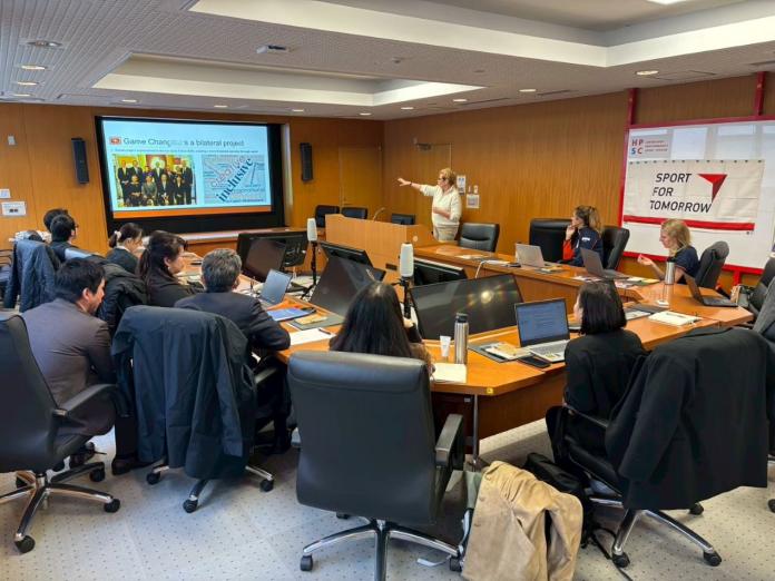 Ten people are sitting around a table, listening to a woman giving a presentation